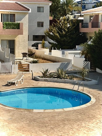 A serene residential area featuring a small, clear blue swimming pool surrounded by a tiled patio. Adjacent to the pool is a white reclining lounge chair. In the background, several two-story houses with red-tiled roofs and plant-lined balconies can be seen. There is also a small set of outdoor furniture, including a table and chairs, and various green plants and trees surround the area.