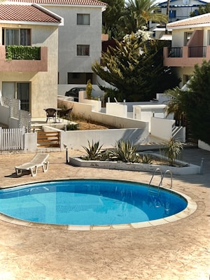A serene residential area featuring a small, clear blue swimming pool surrounded by a tiled patio. Adjacent to the pool is a white reclining lounge chair. In the background, several two-story houses with red-tiled roofs and plant-lined balconies can be seen. There is also a small set of outdoor furniture, including a table and chairs, and various green plants and trees surround the area.