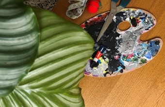 A close-up view of an artist's palette covered with various shades of paint is placed on a wooden surface. Beside it, a pair of scissors and two tubes of paint are visible. A green striped leaf partially covers a portion of the image, adding a touch of nature to the scene.