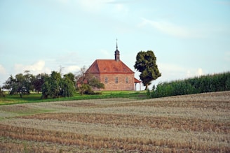 A small, rustic church with a steeple sits surrounded by lush green trees and fields. The building features a red-tiled roof and simple brick walls, creating a serene and peaceful rural scene under a partly cloudy sky.