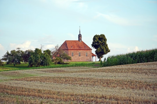 A small, rustic church with a steeple sits surrounded by lush green trees and fields. The building features a red-tiled roof and simple brick walls, creating a serene and peaceful rural scene under a partly cloudy sky.