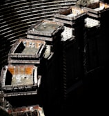 A large construction site with several elevated, rectangular concrete structures surrounded by scaffolding. Workers in safety gear are visible on the platforms. The background shows curved, tiered earthworks suggesting a large excavation or dam. There are shadows that cast a dramatic contrast over the scene.