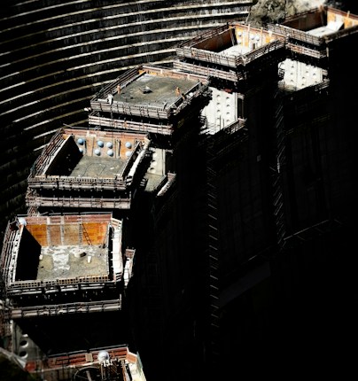 A large construction site with several elevated, rectangular concrete structures surrounded by scaffolding. Workers in safety gear are visible on the platforms. The background shows curved, tiered earthworks suggesting a large excavation or dam. There are shadows that cast a dramatic contrast over the scene.