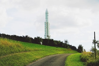 A large white rocket stands vertically in the distance, surrounded by lush green grass and trees. A narrow, curved asphalt path leads towards the rocket, giving the scene a serene and grounded feel.
