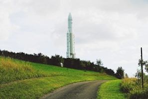 A large white rocket stands vertically in the distance, surrounded by lush green grass and trees. A narrow, curved asphalt path leads towards the rocket, giving the scene a serene and grounded feel.