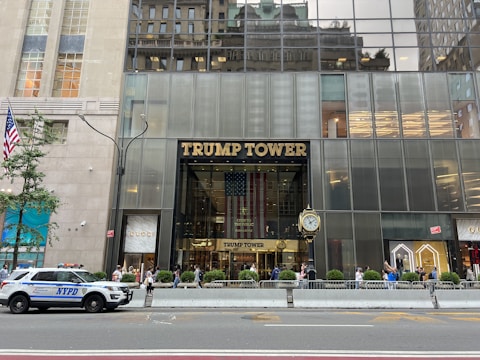 The entrance of a tall building with a glass facade labeled 'Trump Tower' in large gold letters. An American flag is prominently displayed behind the entrance. People are walking on the sidewalk in front, with a police vehicle parked nearby. The surroundings include high-end retail stores and a decorative street clock.
