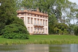 A picturesque, classical-style villa with a light peach facade is surrounded by lush greenery. The two-story building features large windows and ornate details. It's nestled among tall trees beside a tranquil body of water.