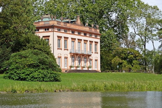A picturesque, classical-style villa with a light peach facade is surrounded by lush greenery. The two-story building features large windows and ornate details. It's nestled among tall trees beside a tranquil body of water.