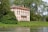 A picturesque, classical-style villa with a light peach facade is surrounded by lush greenery. The two-story building features large windows and ornate details. It's nestled among tall trees beside a tranquil body of water.