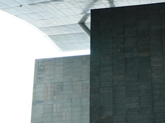 A modern architectural structure with large geometric shapes and a minimalist design. The image features intersecting rectangular and square surfaces composed of gray tiles, with a contrasting white sky in the background.