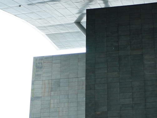 A modern architectural structure with large geometric shapes and a minimalist design. The image features intersecting rectangular and square surfaces composed of gray tiles, with a contrasting white sky in the background.