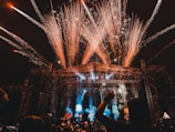 A large outdoor concert or festival with a crowd of people, fireworks exploding above the stage, and colorful lights illuminating the scene. The stage is surrounded by metal scaffolding, and there are various lights and pyrotechnics contributing to the vibrant atmosphere.