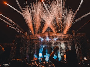A large outdoor concert or festival with a crowd of people, fireworks exploding above the stage, and colorful lights illuminating the scene. The stage is surrounded by metal scaffolding, and there are various lights and pyrotechnics contributing to the vibrant atmosphere.