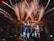 A large outdoor concert or festival with a crowd of people, fireworks exploding above the stage, and colorful lights illuminating the scene. The stage is surrounded by metal scaffolding, and there are various lights and pyrotechnics contributing to the vibrant atmosphere.
