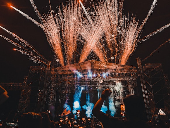 A large outdoor concert or festival with a crowd of people, fireworks exploding above the stage, and colorful lights illuminating the scene. The stage is surrounded by metal scaffolding, and there are various lights and pyrotechnics contributing to the vibrant atmosphere.