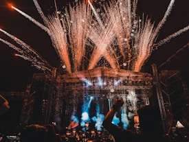 A large outdoor concert or festival with a crowd of people, fireworks exploding above the stage, and colorful lights illuminating the scene. The stage is surrounded by metal scaffolding, and there are various lights and pyrotechnics contributing to the vibrant atmosphere.