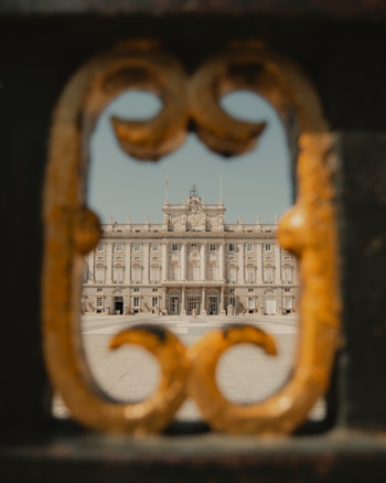 A grand palace is framed through a decorative gate opening, showing intricate architectural details of the building's facade. The ornate metalwork of the gate contrasts with the stately design of the palace.
