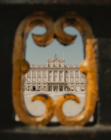 A grand palace is framed through a decorative gate opening, showing intricate architectural details of the building's facade. The ornate metalwork of the gate contrasts with the stately design of the palace.