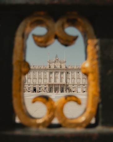 A grand palace is framed through a decorative gate opening, showing intricate architectural details of the building's facade. The ornate metalwork of the gate contrasts with the stately design of the palace.