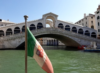 A large stone arch bridge spans across a canal lined with historic buildings. A flag in the foreground features red, white, and green colors. The water below is greenish, and people can be seen walking or standing on the bridge. Bright daylight illuminates the scene, highlighting architectural details.