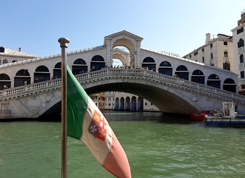 A large stone arch bridge spans across a canal lined with historic buildings. A flag in the foreground features red, white, and green colors. The water below is greenish, and people can be seen walking or standing on the bridge. Bright daylight illuminates the scene, highlighting architectural details.