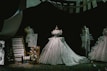 A display of elegant, intricately designed gowns on mannequins, featuring a central dress with a voluminous skirt. The scene includes fashion sketches, fabric swatches, and thread spools, all set against a dark backdrop that highlights the shimmering details of the dresses.