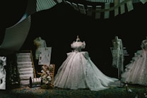 A display of elegant, intricately designed gowns on mannequins, featuring a central dress with a voluminous skirt. The scene includes fashion sketches, fabric swatches, and thread spools, all set against a dark backdrop that highlights the shimmering details of the dresses.