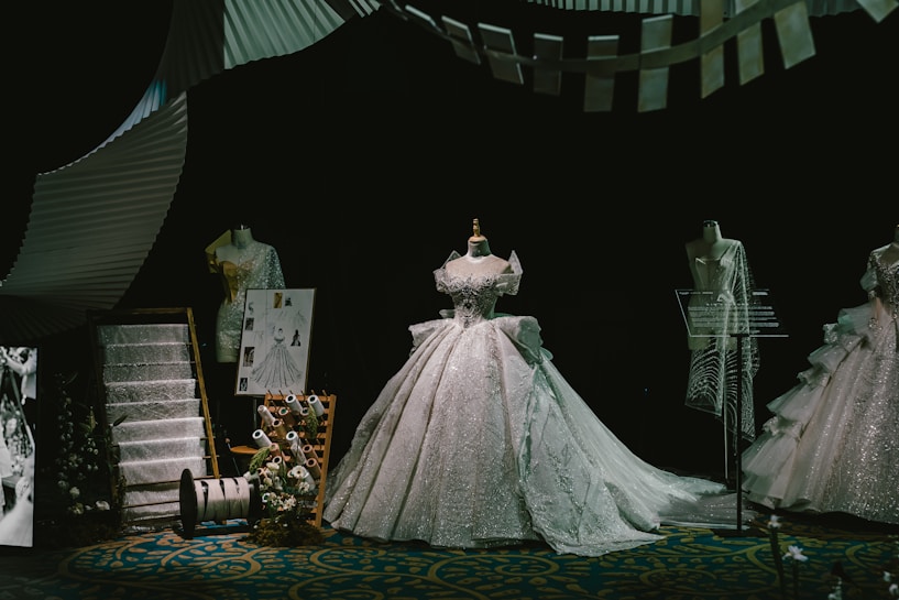 A display of elegant, intricately designed gowns on mannequins, featuring a central dress with a voluminous skirt. The scene includes fashion sketches, fabric swatches, and thread spools, all set against a dark backdrop that highlights the shimmering details of the dresses.