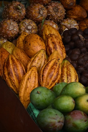 A variety of tropical fruits are displayed in close arrangement, including pineapples, cacao pods, and green mangoes. The fruits are layered in rows, showcasing their distinct colors and textures.
