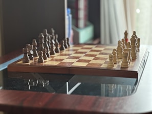 A chessboard is set up on a wooden table, ready for a game. The board is in the standard starting position with pieces placed on both sides, featuring dark and light wooden chess pieces. In the background, there are some blurred items, possibly books or storage boxes, creating a cozy indoor setting.