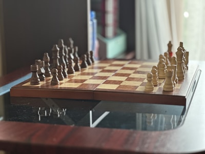 A chessboard is set up on a wooden table, ready for a game. The board is in the standard starting position with pieces placed on both sides, featuring dark and light wooden chess pieces. In the background, there are some blurred items, possibly books or storage boxes, creating a cozy indoor setting.