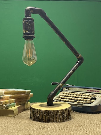 A vintage typewriter is placed on a textured fabric surface beside a stack of hardcover books with colorful spines. In front of the typewriter, there is an industrial-style lamp with an exposed bulb, mounted on a wooden base resembling a tree stump. The background is a solid green color.