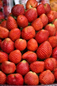 A large pile of fresh, ripe strawberries stacked neatly. The strawberries are vibrant red with tiny seeds covering the surface, and occasional green leaves are visible.
