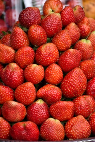 A large pile of fresh, ripe strawberries stacked neatly. The strawberries are vibrant red with tiny seeds covering the surface, and occasional green leaves are visible.