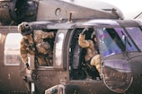 Two soldiers in camouflage uniforms are inside a military helicopter. One soldier is partially outside the open side door, looking outward, while the other is seated inside, gesturing with a raised fist.