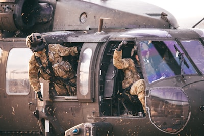 Two soldiers in camouflage uniforms are inside a military helicopter. One soldier is partially outside the open side door, looking outward, while the other is seated inside, gesturing with a raised fist.