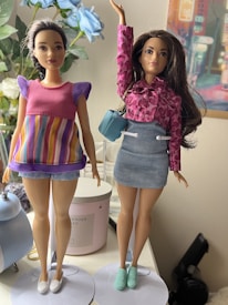 Two dolls are positioned next to each other on a table. The doll on the left is wearing a pink top with purple sleeves and a colorful striped skirt, while the doll on the right is dressed in a pink patterned jacket, a light blue denim skirt, and green shoes. The right doll is holding a light blue handbag. Behind the dolls, there are decorative elements including blue flowers and a colorful wall art piece.