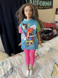 A fashion doll stands dressed in a colorful outfit featuring popular cartoon characters. The doll is wearing a blue top with multicolored sleeves, depicted with animated figures and a cloud pattern. Paired with the top are pink pants and white sneakers. The background includes a faint view of a 'California' poster and various room elements.