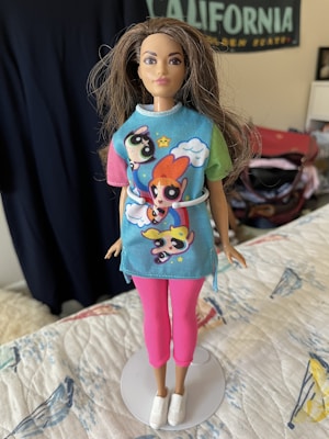 A fashion doll stands dressed in a colorful outfit featuring popular cartoon characters. The doll is wearing a blue top with multicolored sleeves, depicted with animated figures and a cloud pattern. Paired with the top are pink pants and white sneakers. The background includes a faint view of a 'California' poster and various room elements.