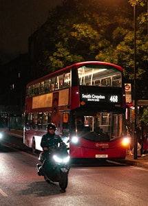 A red double-decker bus is driving down a dimly lit street at night, displaying the route number 468 to South Croydon. The bus is partially filled with passengers visible through the windows. A motorcyclist wearing a helmet rides in front of the bus, casting a long shadow on the road. Trees and streetlights add to the ambiance, with one streetlight illuminating a portion of the scene.