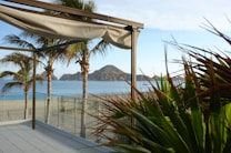 A beautiful beachside scene featuring a clear view of the ocean and distant rocky hills under a clear blue sky. Palm trees frame the foreground, and there is a modern pergola with an unfurled canopy. A glass railing separates the sandy beach from the paved patio, and lush green plants are visible to the right.