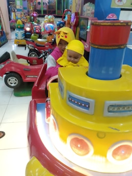 Two children wearing Pikachu-themed outfits sit in a colorful amusement ride surrounded by other vibrant play equipment. The scene is filled with bright colors and playful elements, suggesting a fun and lively environment.