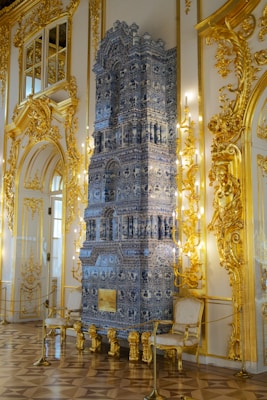 An ornate interior features a highly detailed blue and white ceramic stove that stands prominently between golden elaborately decorated walls. The walls and ceiling are finished with intricate molding and gilded detail, enhancing the luxurious feel of the room. Two elegant chairs with cream upholstery and gilded frames flank the stove. The floor is parquet, complementing the overall opulent design.