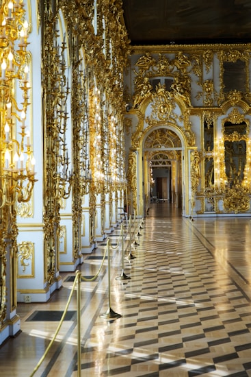 A lavish and ornate hallway featuring intricate gold decorations on the walls and arches, complemented by large mirrors and numerous chandeliers. The parquet flooring adds to the elegance of the space, creating a reflective surface that enhances the light.