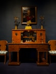 An elegant vintage wooden desk is adorned with ornate gold candelabras. Above it hangs a portrait of a distinguished person in uniform, set against a dark backdrop. Two matching chairs with upholstered seats flank the desk, creating a symmetrical and classical arrangement.