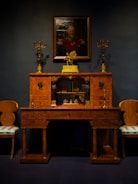 An elegant vintage wooden desk is adorned with ornate gold candelabras. Above it hangs a portrait of a distinguished person in uniform, set against a dark backdrop. Two matching chairs with upholstered seats flank the desk, creating a symmetrical and classical arrangement.
