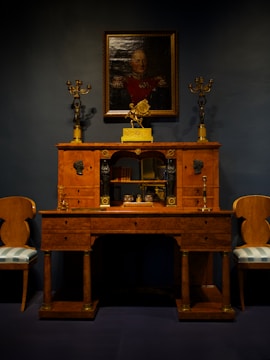 An elegant vintage wooden desk is adorned with ornate gold candelabras. Above it hangs a portrait of a distinguished person in uniform, set against a dark backdrop. Two matching chairs with upholstered seats flank the desk, creating a symmetrical and classical arrangement.