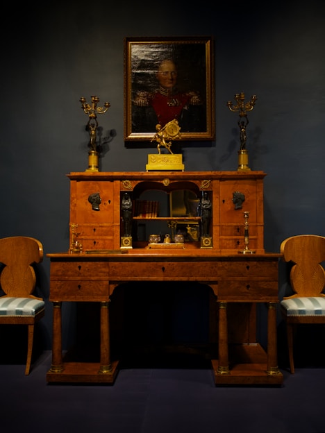 An elegant vintage wooden desk is adorned with ornate gold candelabras. Above it hangs a portrait of a distinguished person in uniform, set against a dark backdrop. Two matching chairs with upholstered seats flank the desk, creating a symmetrical and classical arrangement.