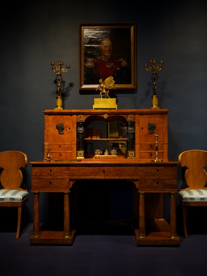 An elegant vintage wooden desk is adorned with ornate gold candelabras. Above it hangs a portrait of a distinguished person in uniform, set against a dark backdrop. Two matching chairs with upholstered seats flank the desk, creating a symmetrical and classical arrangement.
