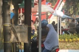 A person wearing a blue helmet is riding a motorcycle with a passenger who is wearing a polka-dotted headscarf. There are trees and a sign in the foreground, with a street scene in the background featuring people, plants, and a Coca-Cola sign.